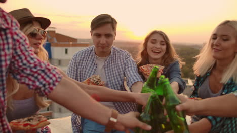 Young-people-clinks-glasses-and-drinks-beer-from-green-bottels-on-the-party-with-friends-on-the-roof-at-the-sunset.They-eat-hot-pizza-after-in-summer-everning.